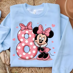 May include: Light blue sweatshirt featuring a pink polka-dotted number 8 and a cartoon character with a pink bow. The character is smiling and wearing a pink dress. Small pink hearts are scattered around the design.