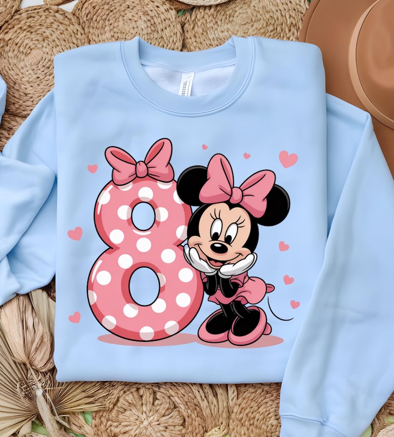 May include: Light blue sweatshirt featuring a pink polka-dotted number 8 and a cartoon character with a pink bow. The character is smiling and wearing a pink dress. Small pink hearts are scattered around the design.