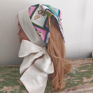 May include: A headscarf with a colorful insect print, featuring pink, teal, and green squares. The scarf has a cream-colored border and a large bow tied under the chin. The head covering is designed to be worn on the head.