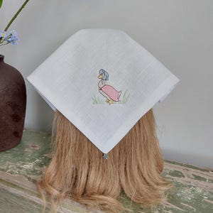 May include: A white square handkerchief with an embroidered duck wearing a bonnet and pink dress. The duck is standing in green grass. A small silver charm hangs from the corner of the handkerchief.