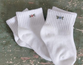 以下が含まれることがあります： 白いアンクルソックス2足組。リブ編みの履き口に、ピンク、青、緑色の小さな花の刺繍が施されています。靴下は柔らかい素材、おそらく綿で作られています。