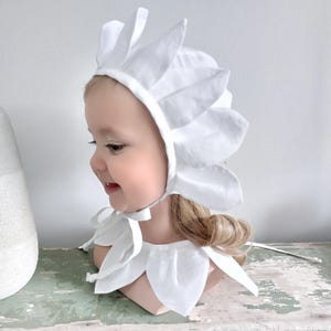 Peut inclure: Ensemble bonnet et col pour bébé de couleur blanche. Le bonnet présente des formes en forme de pétales autour de la tête et un lien sous le menton. Le col assorti a des détails similaires en forme de pétales. L'ensemble est en tissu blanc doux.