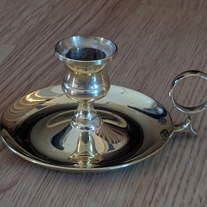 May include: A brass candlestick holder with a circular base and a handle. The candle holder has a cup-shaped top and a decorative stem. The handle is a small ring. The candlestick holder is on a wooden surface.
