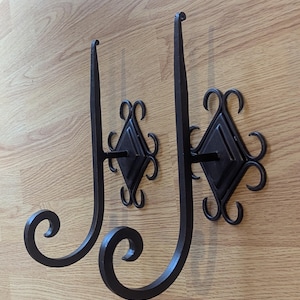 May include: Two wrought iron wall sconces. Each sconce features a diamond-shaped backplate with decorative scrollwork and a long, curved arm ending in a scroll. The sconces are dark brown and mounted on a light brown wooden surface.