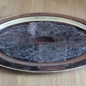 May include: Oval silver serving tray with ornate, engraved floral and shell design. The tray has two curved handles and a polished finish. A decorative piece for serving food or displaying items.