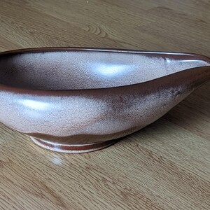 May include: A ceramic gravy boat with a unique shape and a handle. The interior is a light beige color, while the exterior rim and handle are a darker brown. The boat sits on a small, circular base.
