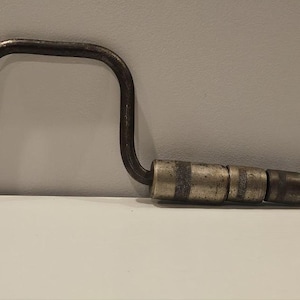 May include: An antique hand drill with a dark metal frame and a wooden handle. The drill has a curved handle and a circular knob on one end. The handle is made of a series of metal and wooden rings.