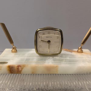 May include: A vintage desk clock and pen holder set. The clock is square with a gold-tone frame and a cream-colored face. Two gold-tone pen holders flank the clock on a rectangular marble base with brown and white veining. The clock face reads "SCOTTCO".