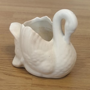 May include: White ceramic swan planter with a textured surface. The planter is shaped like a swan with its head turned to the side. The swan's neck is curved and its beak is pointed. The planter has a small opening at the top for planting.