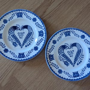 May include: Two white ceramic plates with a blue floral border and a central design of two birds forming a heart shape. The birds are blue with intricate patterns. The plates are on a wooden surface.