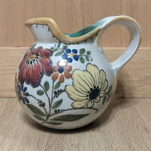 May include: A ceramic pitcher with a round body and a curved handle. The pitcher is decorated with colorful floral designs in red, yellow, blue, and brown. The rim and handle have a light brown trim.