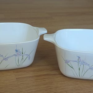 May include: Two off-white, square-shaped casserole dishes with handles. Each dish features a floral design of purple irises and green stems. The dishes are set on a wooden surface.