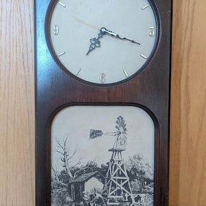 May include: A rectangular, dark wood-framed wall clock. The top section features a round clock face with black hands and numerals. The bottom section displays a black and white rural scene with a windmill and cows.