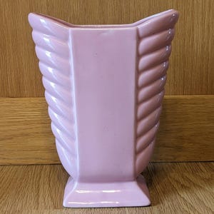 May include: A pink ceramic vase with a unique, twisted design. The vase has a square base and a wide opening.