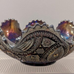 May include: An iridescent glass bowl with a scalloped rim and intricate etched patterns. The bowl's interior displays a blend of gold, purple, and blue hues. The decorative bowl sits on a clear, ridged surface.
