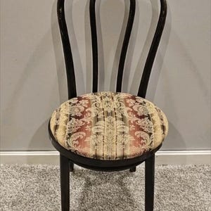 May include: Dark brown wooden chair with a round seat and curved back. The seat features a patterned fabric in shades of red, gold, and cream. The chair has four legs and is set against a neutral background.