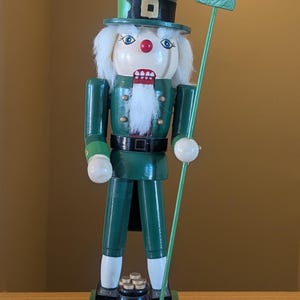 May include: A green wooden nutcracker in the shape of a leprechaun, wearing a top hat with a shamrock, holding a staff with a shamrock, and standing on a green base with a pot of gold. The nutcracker has a white beard and a red mouth.