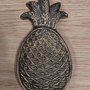 May include: A silver-colored pineapple-shaped trinket dish. The dish has a detailed textured surface, with a pattern of diamond shapes. The pineapple's crown is also detailed with textured leaves. The dish is placed on a wooden surface.