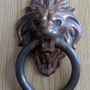 May include: Bronze lion head door knocker with a circular handle. The lion's face is detailed with a mane and facial features. The knocker is mounted on a wooden surface.
