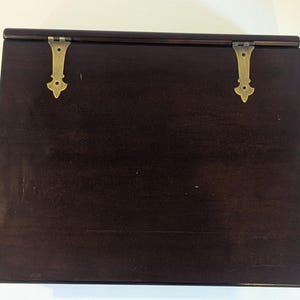 May include: A dark brown wooden box with a hinged lid. The lid is secured by two brass-colored hinges. The box has a smooth, polished surface, and the wood grain is visible. The box is rectangular and appears to be empty.