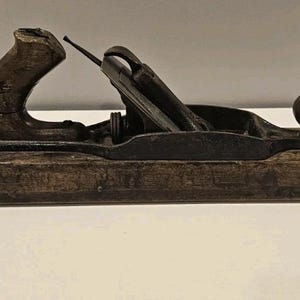 May include: A vintage wooden hand plane with a dark metal blade and a wooden handle. The plane has a rectangular base and a curved handle on one end. The handle is worn, showing signs of age and use. The plane is a woodworking tool.