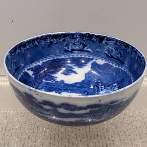 May include: A blue and white ceramic bowl with a landscape design. The bowl features a detailed pattern of buildings, trees, and clouds. The rim has a detailed border. The bowl is a classic piece of tableware.