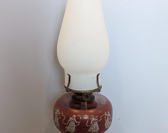 May include: An antique oil lamp with a clear glass base and a frosted white glass shade. The lamp's red base features a white relief design of dancing figures. The lamp has brass accents and a clear glass chimney.