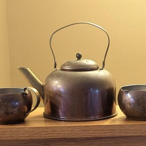 May include: A vintage tea set with a copper finish. The set includes a teapot with a curved handle, a lid, and a spout, along with two small cups with handles. The tea set is displayed on a wooden surface.