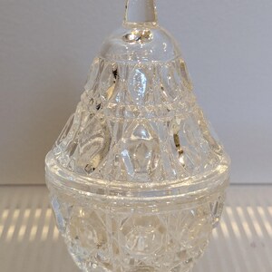 May include: A clear, faceted glass jar with a lid. The jar has a rounded base and a conical lid with a small knob on top. The glass has a diamond-like pattern, reflecting light. The jar is likely for storing small items or as a decorative piece.