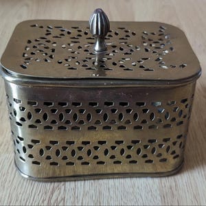 May include: A vintage brass box with a decorative pierced design on the lid and sides. The box has a square shape with rounded corners and a small, ornate handle on top. The brass has a slightly aged patina, giving it a classic look.