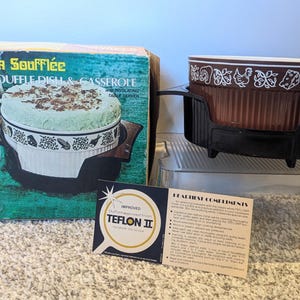 May include: A vintage La Soufflée souffle dish and casserole set. The brown and white dish sits in a black holder. The box features the words "La Soufflée" and "Nordic Ware". A Teflon II sticker is also visible.
