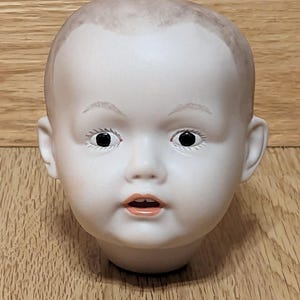 May include: A vintage porcelain doll head with a pale complexion, black eyes, and a slightly open mouth. The doll head has a bald head with a hint of painted hair and is set against a wooden background. The doll head is a collectible item.