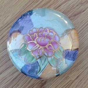 May include: A round, decorative ceramic piece with a floral design. The center features a pink peony with gold outlines and green leaves. The background has a mix of colors including blue, purple, and gold.