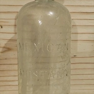 May include: Clear glass bottle with embossed text reading "MEXICAN MUSTANG LINIMENT." The bottle has a rounded body and a narrow neck. The background is a light brown wooden surface.