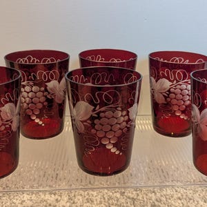 May include: Set of six ruby red drinking glasses. Each glass features a white etched design of grapes and vines. The glasses are cylindrical with a slightly flared top and are displayed on a clear shelf.