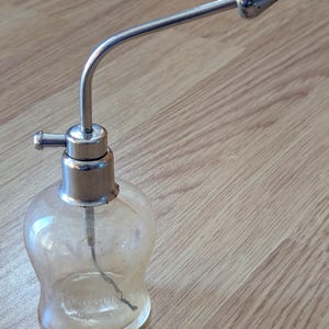 May include: A vintage clear glass perfume atomizer with a silver-toned metal spray nozzle and pump. The bottle has a rounded shape and sits on a wooden surface. The atomizer is likely from the early to mid-20th century.