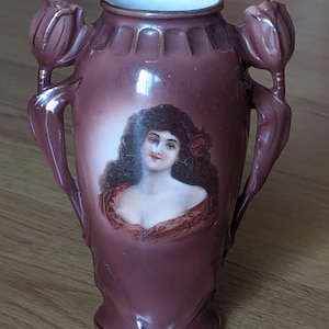 May include: A maroon-colored ceramic vase with a portrait of a woman. The vase has two handles shaped like tulip buds. The woman in the portrait has dark curly hair and a red rose. Gold trim accents the rim and base.