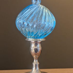 May include: A clear, light blue glass bottle with a bulbous, swirled design sits atop a silver-colored metal candlestick. The candlestick has a round base and a slender, ornate stem.