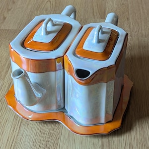 May include: A white and orange ceramic tea set with a teapot and creamer, resting on a matching tray. The set features a geometric design with orange trim and handles. The set is designed for serving tea.