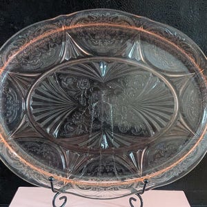 May include: Pink Depression glass oval serving platter with a detailed floral and geometric pattern. The platter has a scalloped edge and is displayed on a black metal stand. The glass has a subtle pink hue and intricate embossed designs.