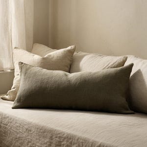 May include: A neutral-toned bedroom scene featuring a bed with several pillows. A long, olive-green rectangular pillow rests on top of the bed, with other pillows in varying shades of beige. A window with sheer white curtains is visible.