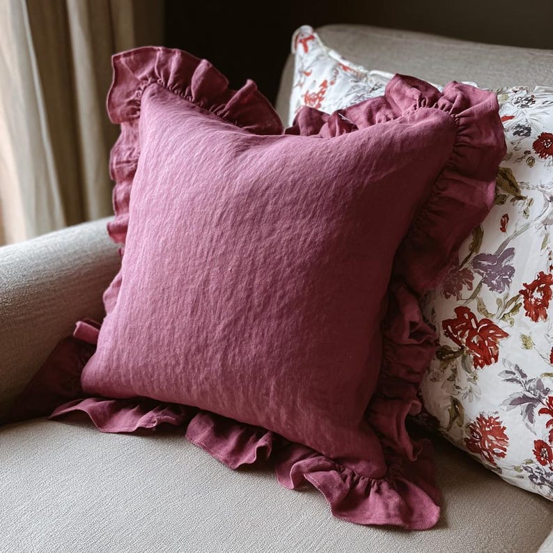 May include: A square cushion in a deep rose hue with a ruffled trim. The cushion has a textured appearance and is placed on a neutral-toned sofa. A floral patterned cushion is partially visible in the background.