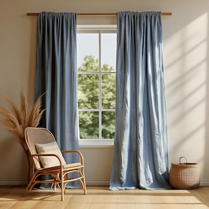 May include: Two light blue linen curtains hang from a wooden rod, framing a window with a view of green trees. A wicker chair with a cream-colored pillow sits to the left, and a woven basket is on the right.