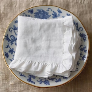 May include: A stack of white linen napkins with ruffled edges, resting on a blue and white patterned plate with a gold rim. The plate features a floral design.