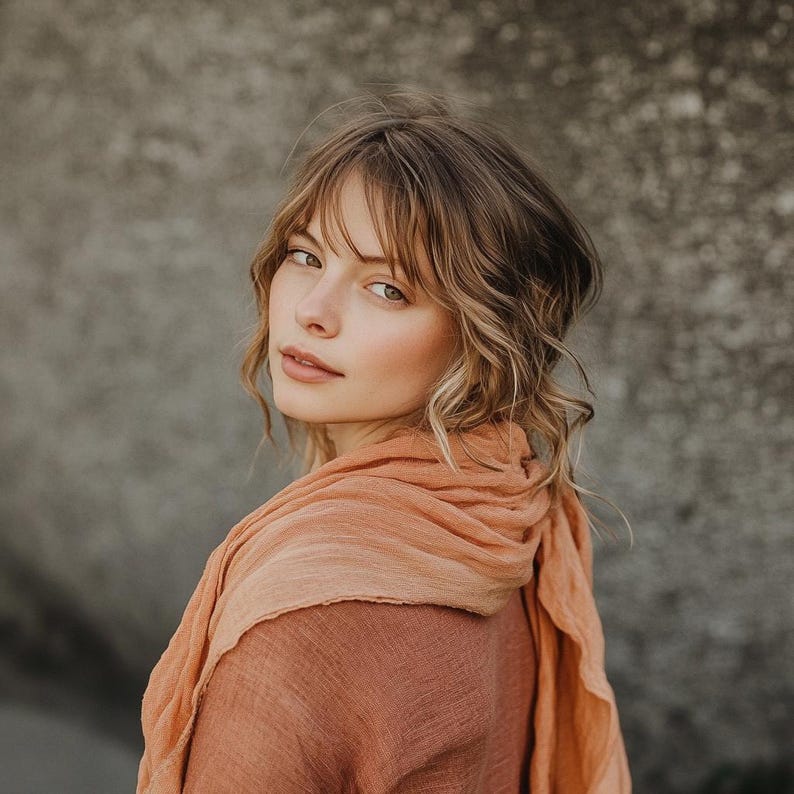 May include: A woman with light brown hair and a peach-colored scarf. The scarf is draped around the neck and shoulders, with a gradient from a darker peach to a lighter shade. The woman is looking over her shoulder at the viewer.