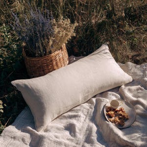 Op de afbeelding: Een lang, rechthoekig, lichtbeige linnen kussen ligt op een geweven deken. Een rieten mand gevuld met gedroogde bloemen en een bord met koekjes en een kopje staan ​​in de buurt, wat een picknick suggereert.