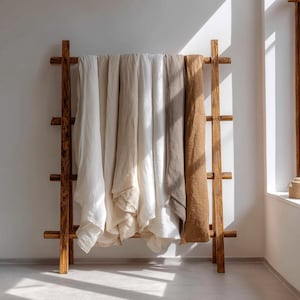 May include: A wooden ladder-style rack displays folded linen textiles in shades of white, beige, and brown. The rack is made of natural wood and stands against a white wall, with sunlight streaming in from a nearby window.