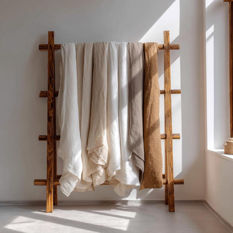 May include: A wooden ladder-style rack displays folded linen textiles in shades of white, beige, and brown. The rack is made of natural wood and stands against a white wall, with sunlight streaming in from a nearby window.