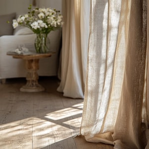May include: Beige linen curtains hang in front of a window, with sunlight streaming through. A small wooden side table holds a vase of white flowers. The floor is made of light-colored wood.