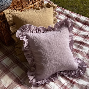 May include: Two square cushions with frilled edges. One cushion is a light lavender colour, and the other is a tan colour. The cushions are on a red and white plaid blanket, with a wicker basket in the background.
